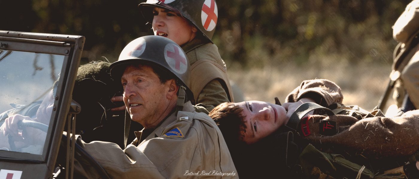 A young soldier sits on the back of a military ambulance, looking contemplative and weary. He wears a standard-issue uniform with a helmet slightly askew. The ambulance, marked with a red cross, is parked on a dusty road with hints of a battlefield in the background. The soldier’s expression reflects a mix of determination and vulnerability as he takes a moment to rest. The scene captures the essence of the challenges faced by soldiers during wartime, highlighting both their strength and the toll of conflict.